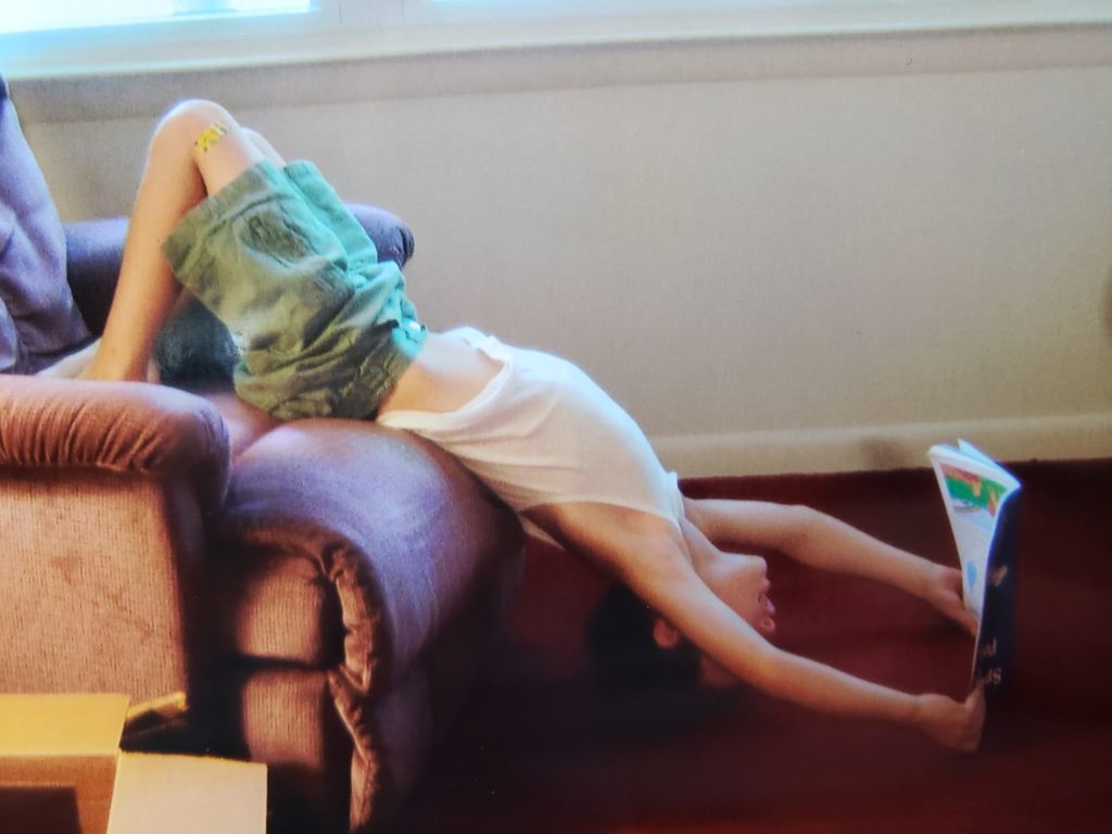 young boy reading book while on back leaning off of chair with head on floor