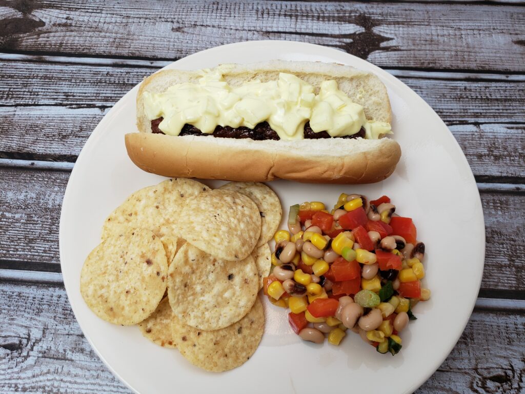 A plate with hot dog with onion relish, tortilla chips and corn salsa.