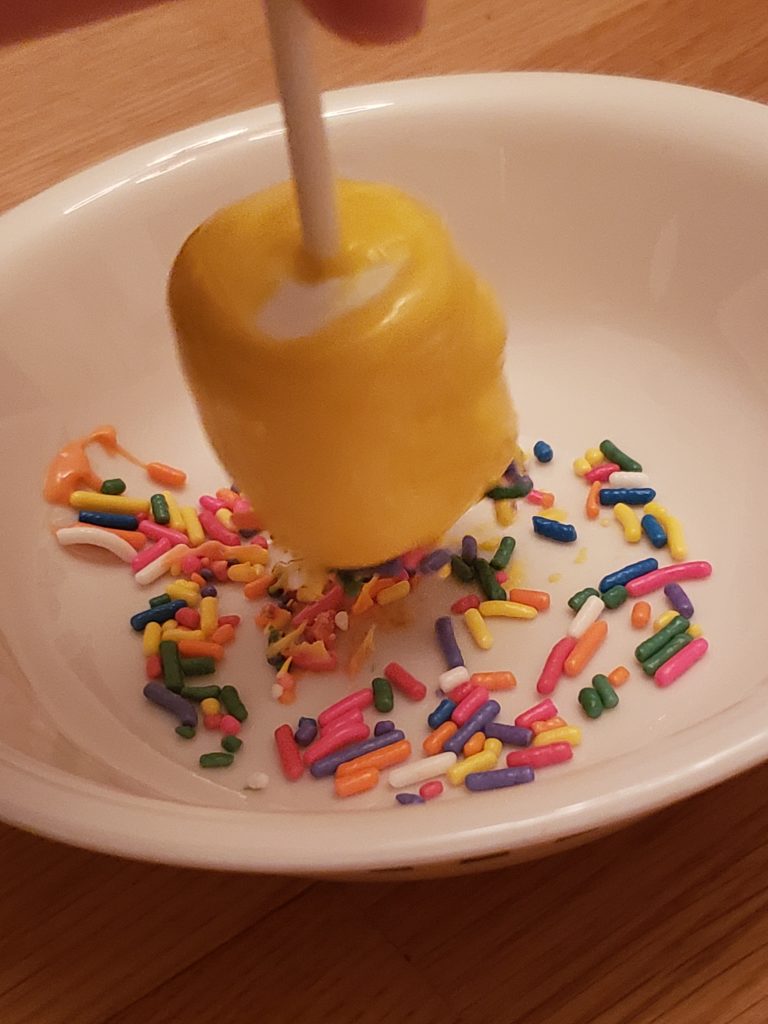 dipping yellow Marshmallow Pop in colored sprinkles