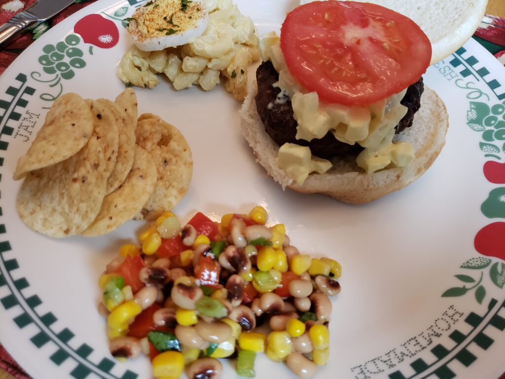 hamburger on a plate with tortilla chips, macaroni salad and corn and red pepper salsa