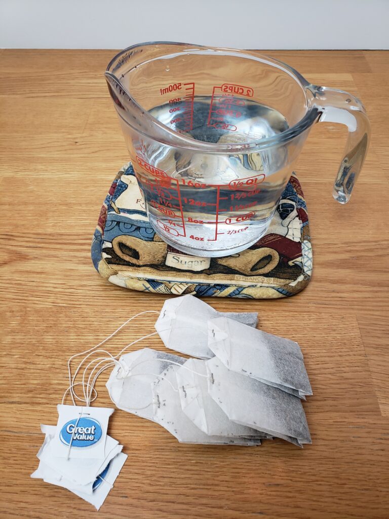 boiling water in Pyrex cup on pot holder and six tea bags lying in front of it.