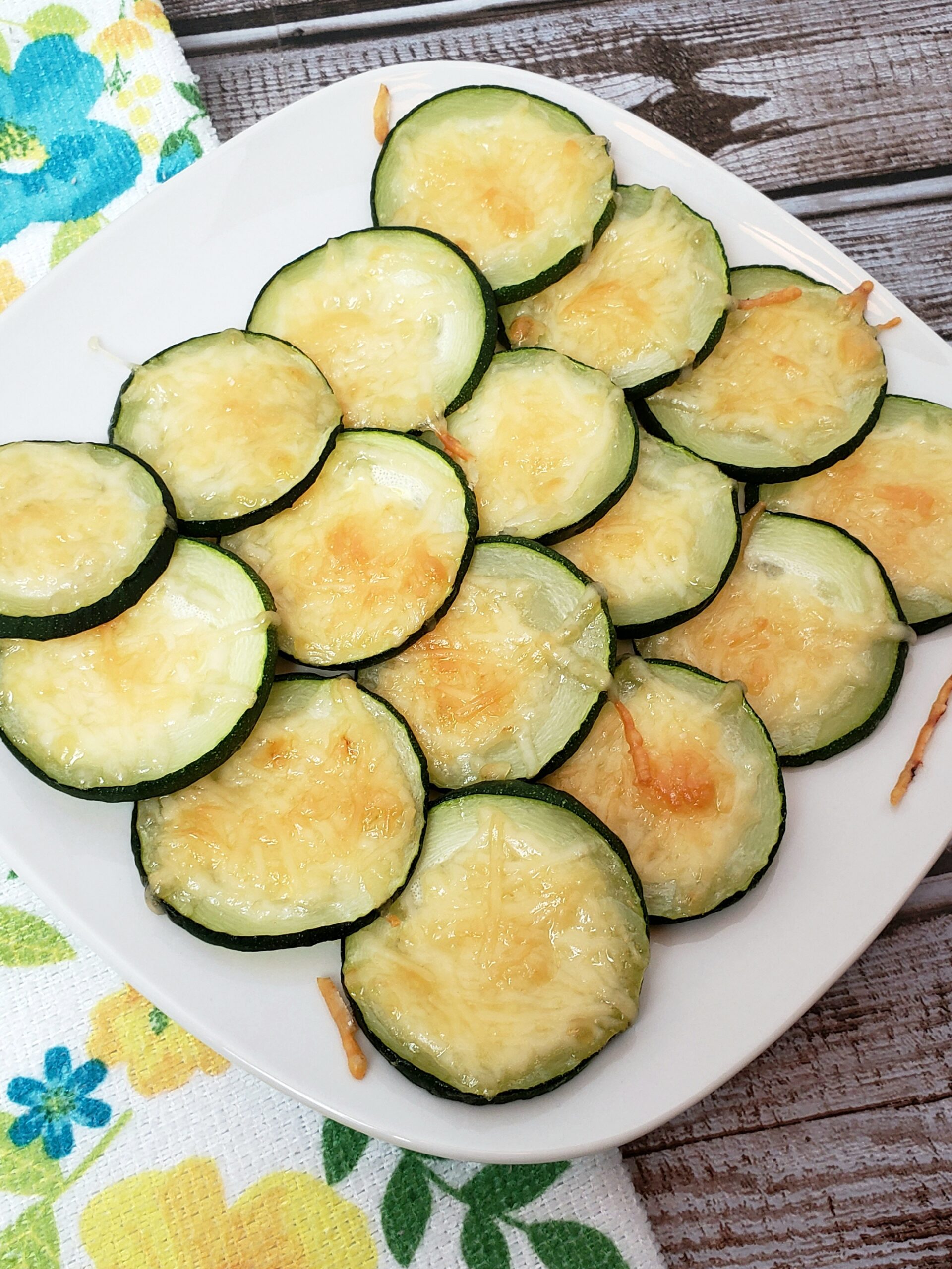 Cheesy Zucchini Coins on white plate next to kitchen towel with flowers
