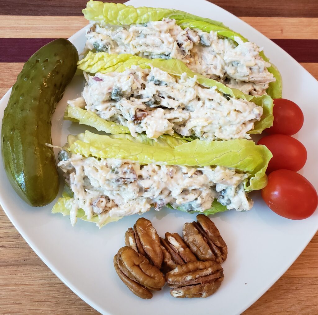 deluxe Keto chicken salad stuffed into romaine lettuce leaves and on a white plate with a pickle, 3 cherry tomatoes and 5 pecan halves.