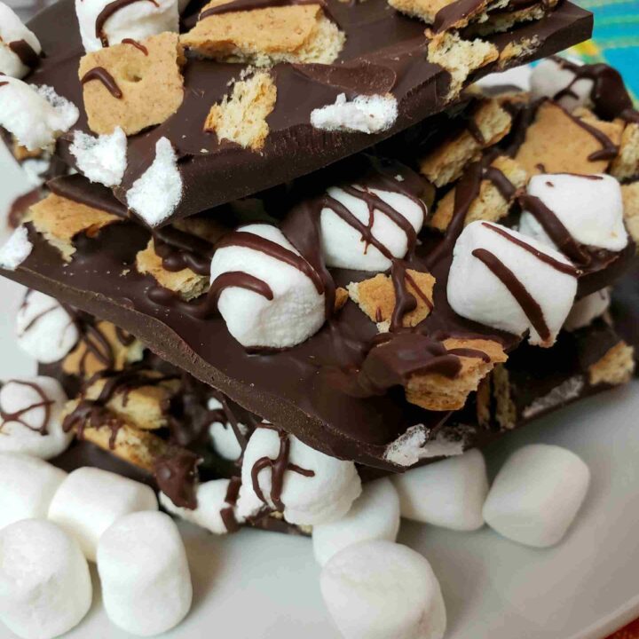 s'mores bark broken and stacked on a white plate with mini marshmallows in front of it.