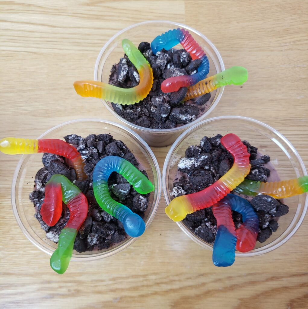 chocolate pudding in 3 plastic cups with 3 gummy worms each and crushed Oreos on top